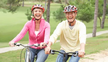 Two smiling people wearing cycling helmets are riding bicycles together in a park-like setting.