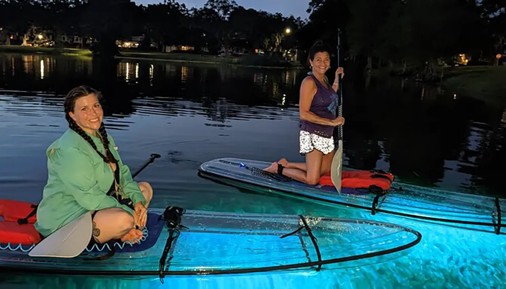 Two people are kneeling on illuminated stand-up paddleboards on a calm body of water during twilight