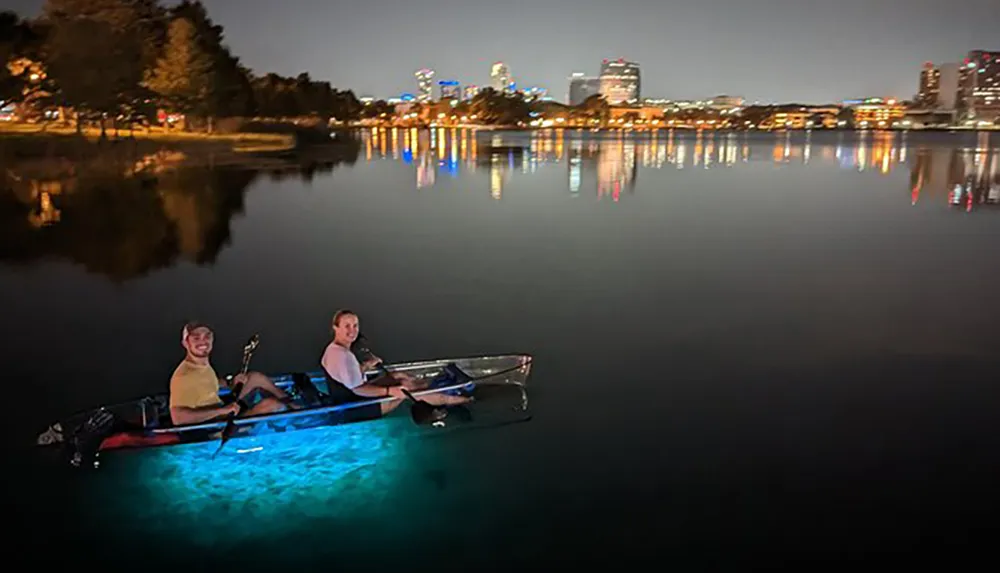 Two people are night kayaking on a calm water body with colorful underwater lights illuminating their path and a city skyline in the background