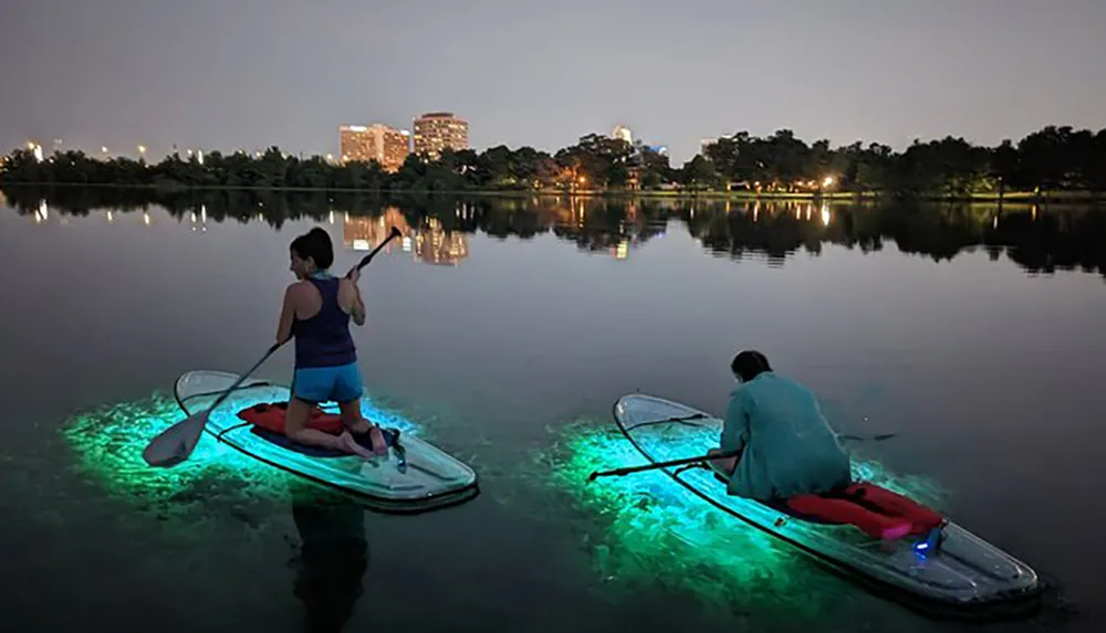Two people are paddleboarding at night on a calm lake illuminated by lights underneath their boards with a city skyline in the background