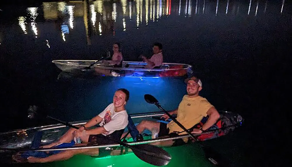 Two pairs of people enjoy nighttime kayaking with vibrant underwater lights illuminating the water around their clear kayaks