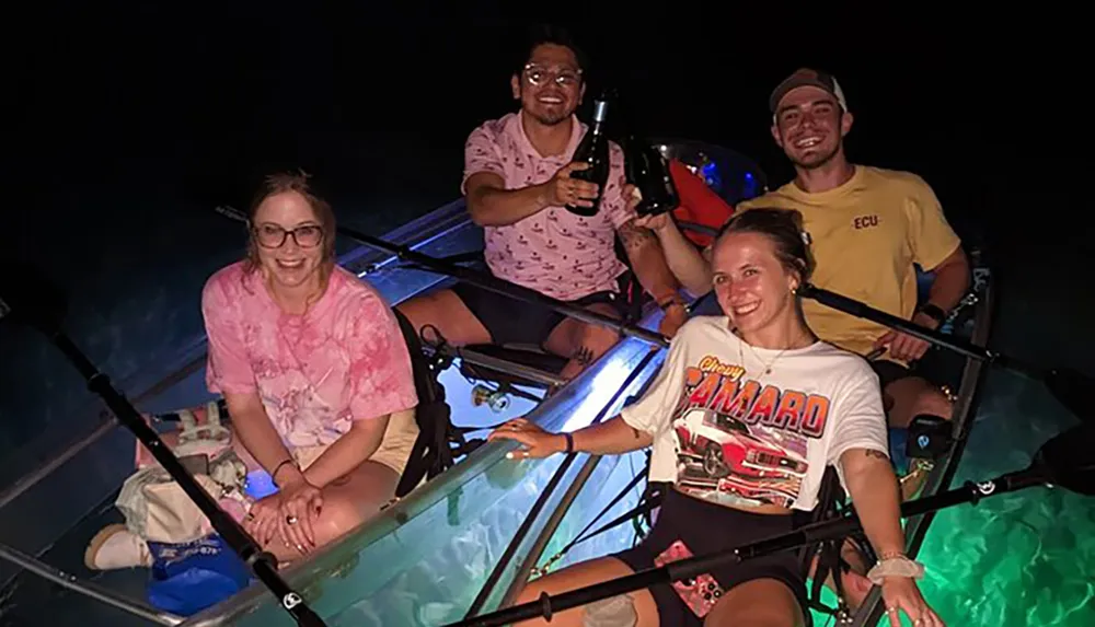 Four people are smiling and posing at night on a transparent canoe with underwater lighting that reveals the sea life below
