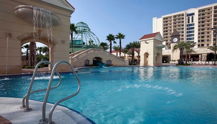 The image shows a luxurious outdoor swimming pool area with water features palm trees and a hotel building in the background under a clear sky