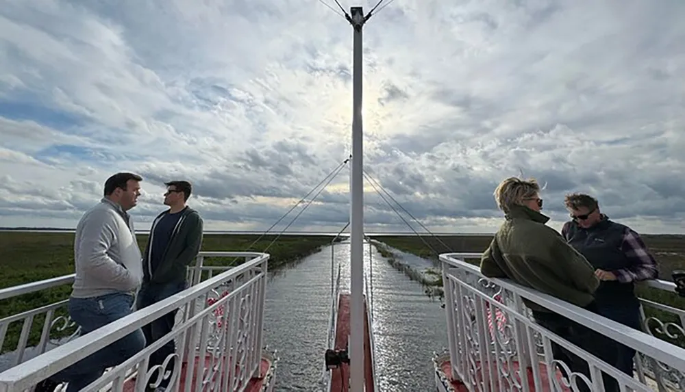 A group of people are having conversations on a narrow bridge with ornate railings extending over a flat landscape under a dramatic sky