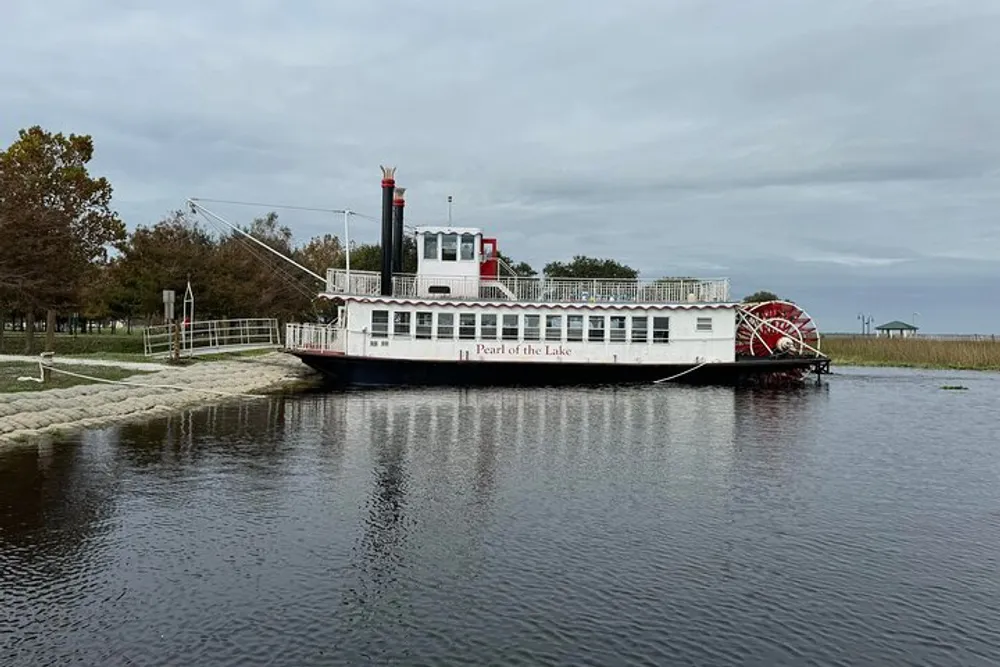 A paddlewheel boat named Pearl of the Lake is docked at a calm lakeside with an overcast sky above