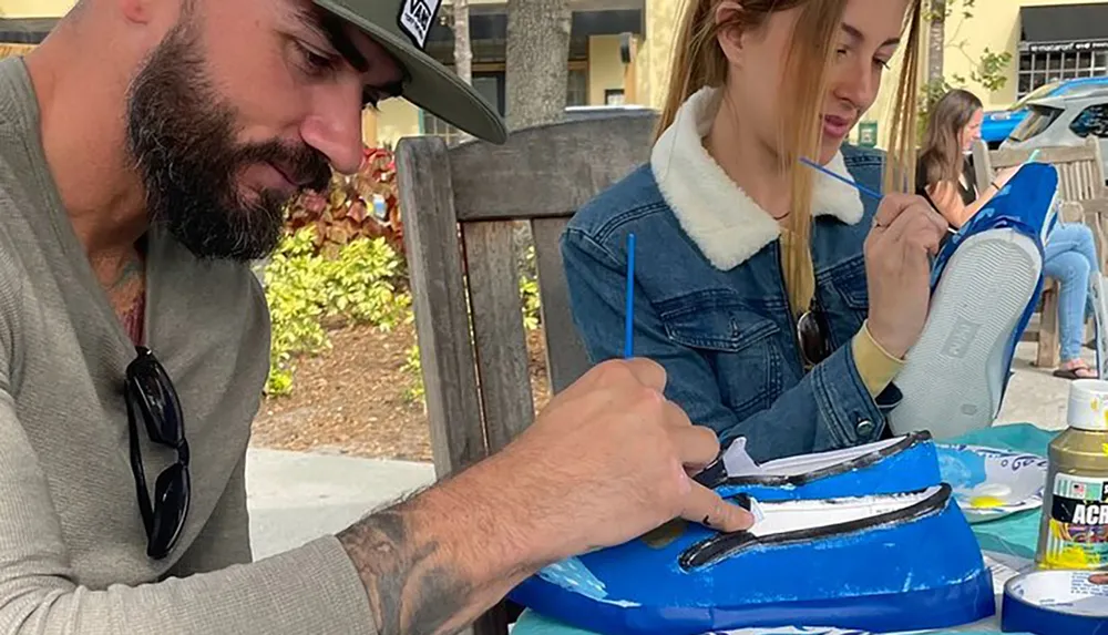 Two people are intently painting sneakers at an outdoor crafting table