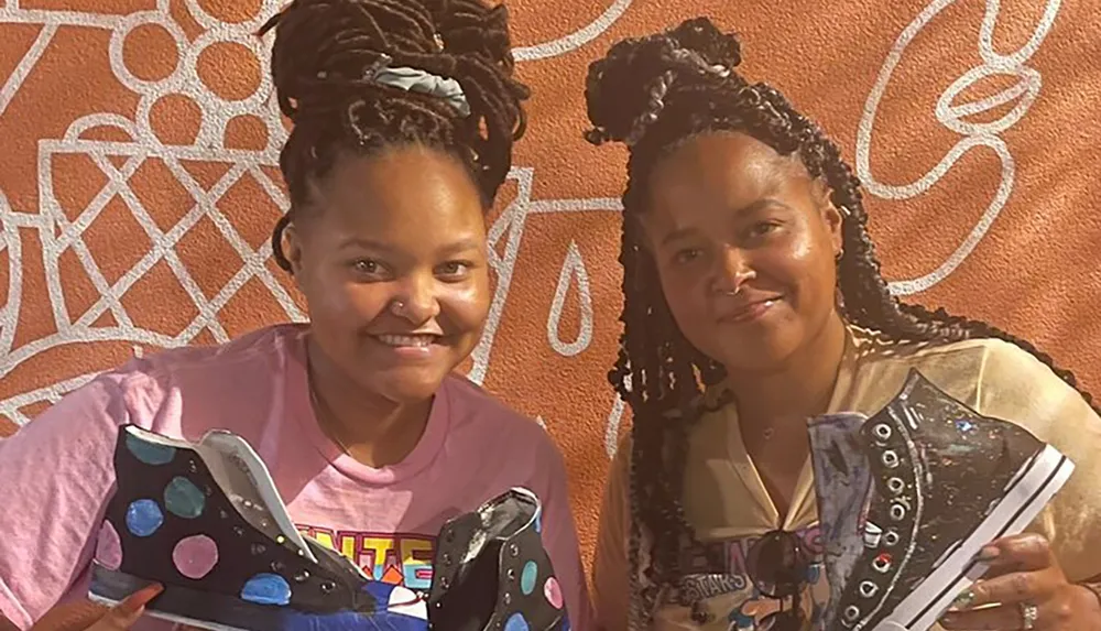 Two individuals are smiling for the camera while holding decorated sneakers standing in front of an orange wall with white line patterns