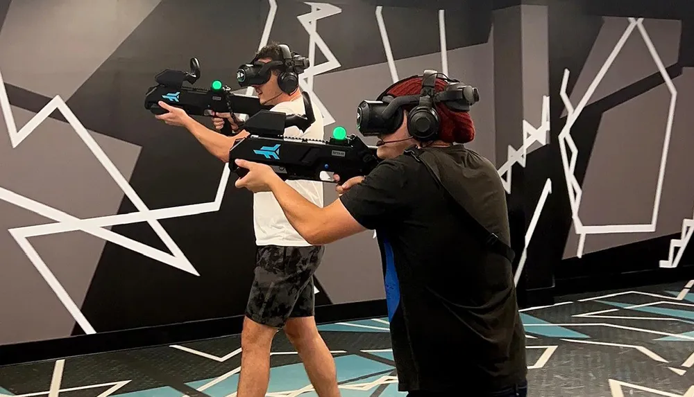 Two people are immersed in a virtual reality experience wielding VR guns and wearing headsets in an indoor space with abstract patterns on the walls