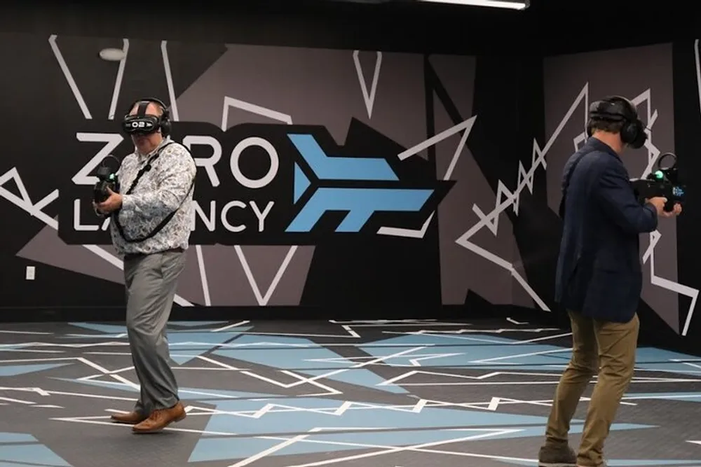 Two people are immersed in a virtual reality experience in a room with dynamic graphic patterns and the text ZERO LATENCY on the wall