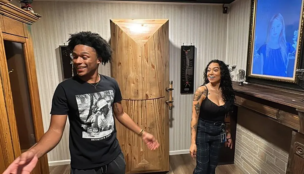 A man and a woman are smiling and posing in a room with a rustic wooden door and a painted portrait on the wall