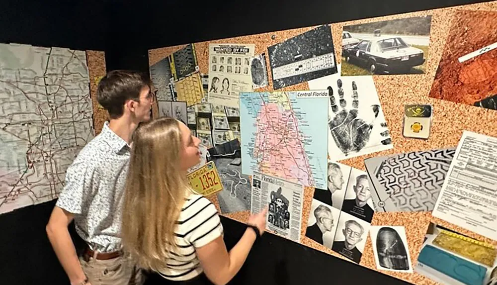 Two individuals are examining a display board covered with various items including maps documents and photographs