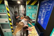 A group of people are engaging in what appears to be an escape room challenge, surrounded by themed decorations that simulate a technical or possibly spacecraft environment.