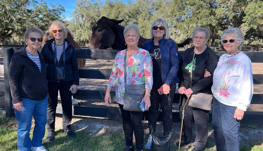 A group of smiling women poses for a photo in front of a fenced area with a horse peeking its head over the fence behind them.
