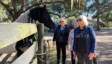 A black and white horse peers over a wooden fence at three smiling women in a sunny, tree-filled setting.