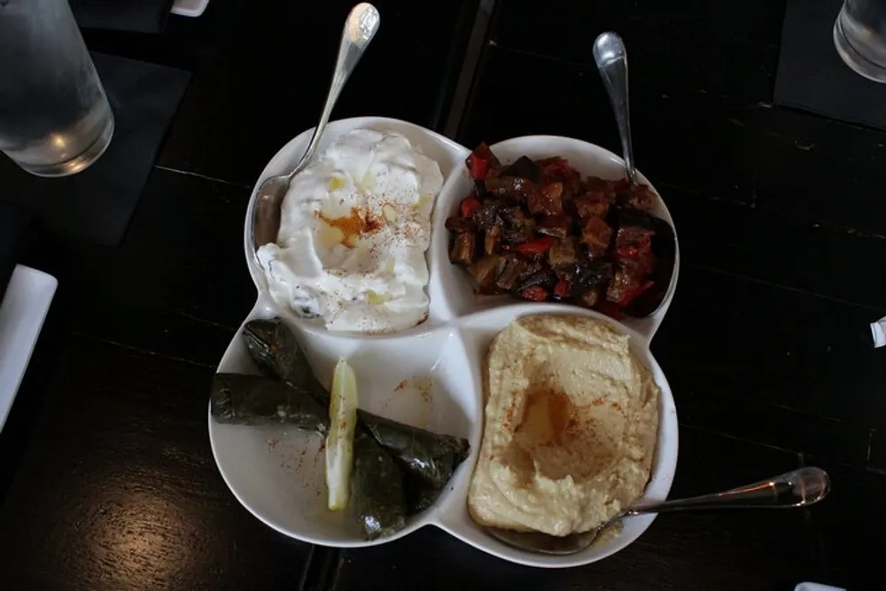 This image shows a dish with four compartments containing hummus yogurt roasted vegetables and stuffed grape leaves commonly found in Middle Eastern cuisine