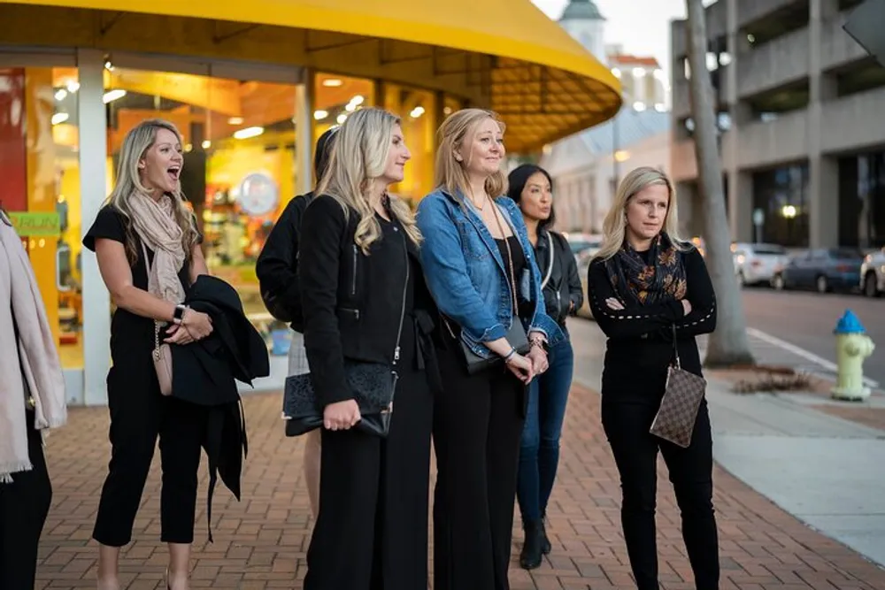 A group of women dressed in casual to smart-casual attire stand on a sidewalk looking to their left with various expressions ranging from amusement to curiosity