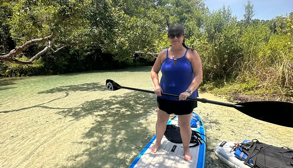 A person stands on a paddleboard in shallow clear water near greenery with another board alongside