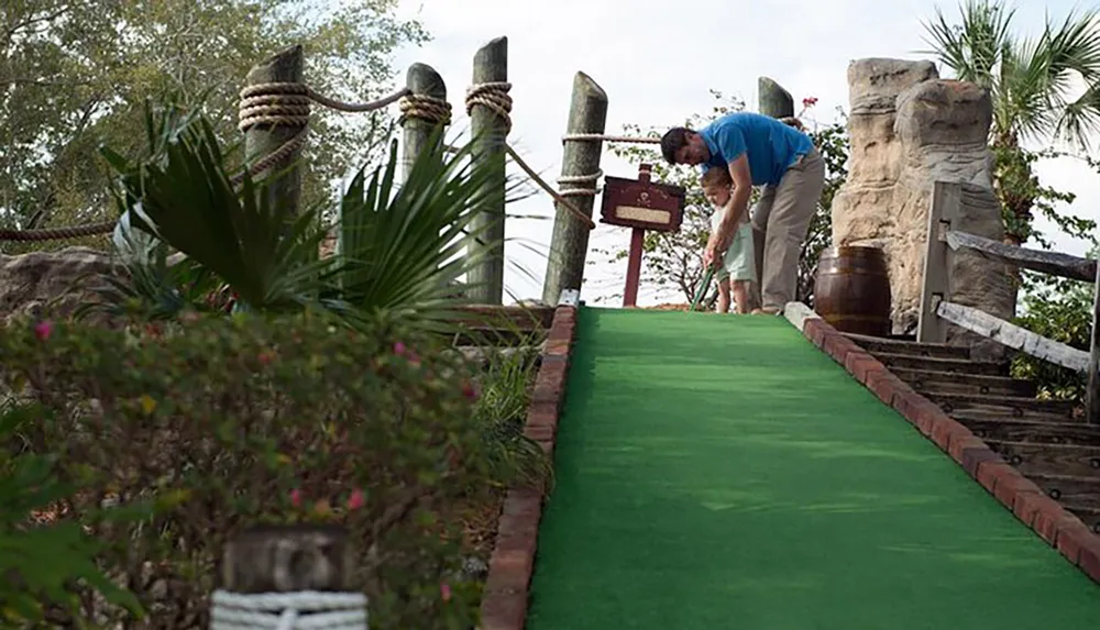 An adult is teaching a young child how to putt on a pirate-themed miniature golf course