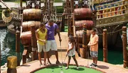 A family of four is enjoying a game of miniature golf with a pirate ship backdrop.