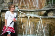 A young girl with a sunny smile holds onto the ropes of a rope bridge in front of a softly blurred waterfall background.
