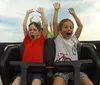 Two children are exuberantly raising their arms and yelling with joy while riding a roller coaster