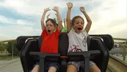 Two children are exuberantly raising their arms and yelling with joy while riding a roller coaster.
