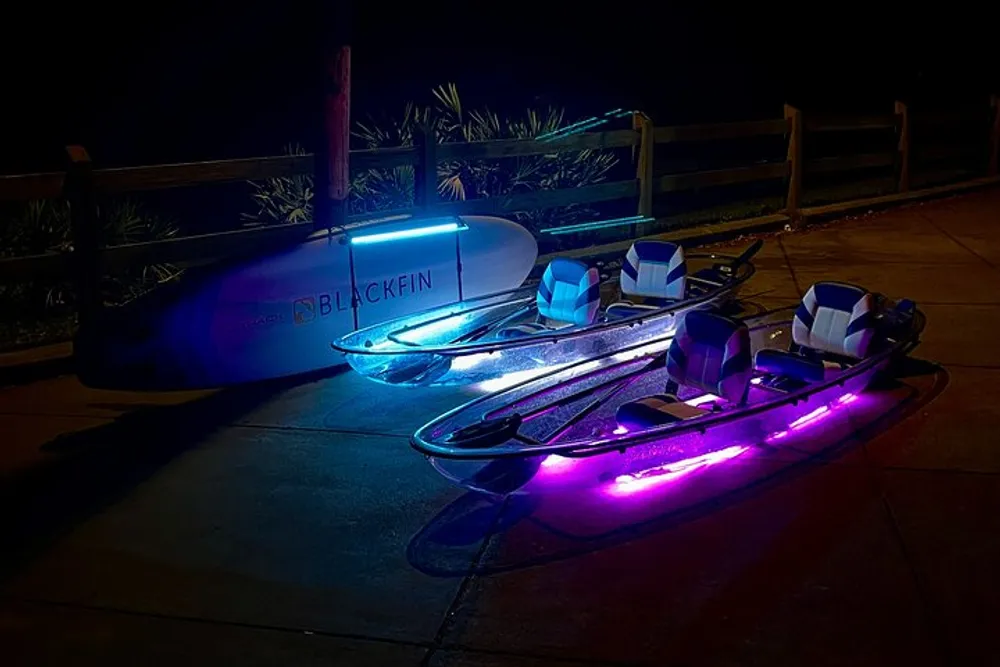 Two illuminated paddleboards with neon lights are lying on a pavement at night next to an upright paddleboard named BLACKFIN