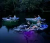 People are kayaking at night with their vessels illuminated by bright colorful underwater lights