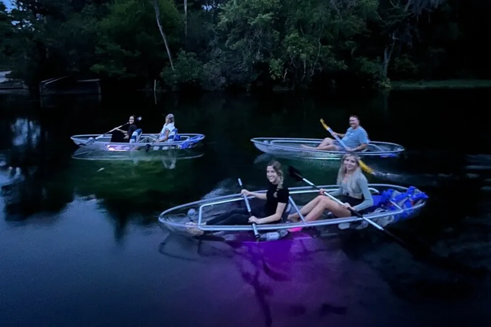 Four people are sitting in clear kayaks with colored lights on a body of water during dusk surrounded by trees