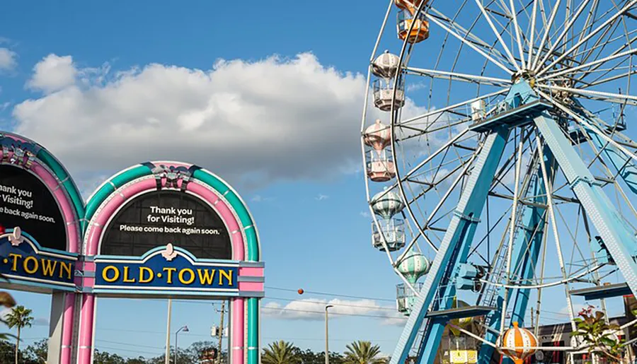 The image shows a colorful entrance sign for OLD TOWN with a thank you message and a Ferris wheel against a partly cloudy sky.