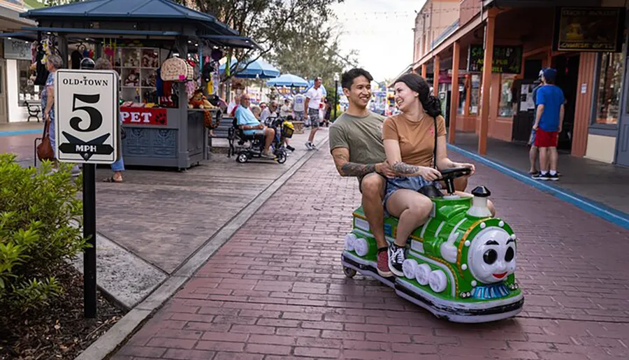 Two people share a light moment as they ride on a whimsical train-shaped scooter down a pedestrian street, passing by a speed limit sign indicating 5 mph in an area called Old Town.