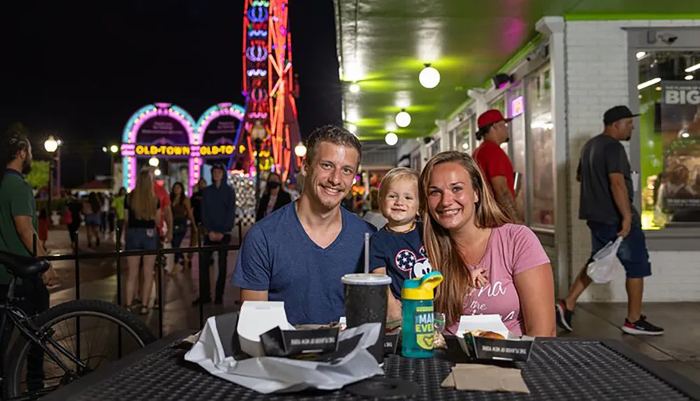 A family of three is smiling at the camera while sitting at an outdoor table with food in a lively amusement park at night