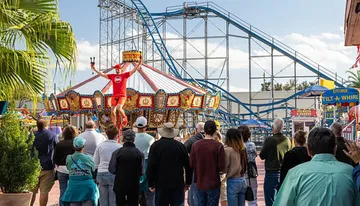 A crowd is watching a performer who is balancing on a unicycle and juggling clubs at an amusement park, with a carousel and rollercoaster in the background.