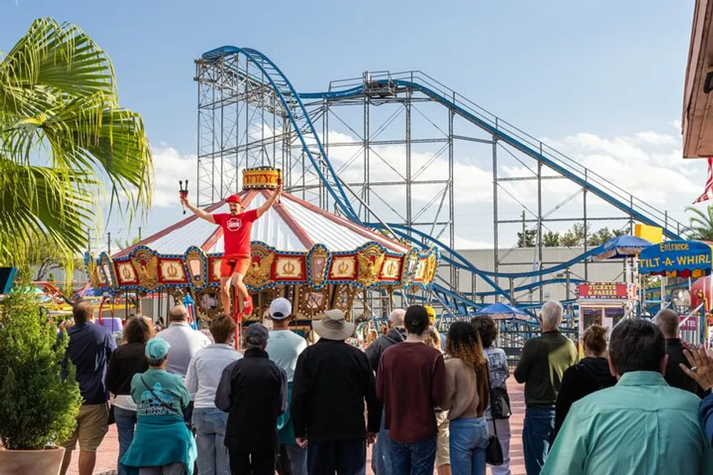 A crowd of people watches a performer on stilts at an amusement park with a merry-go-round in the foreground and a roller coaster in the background