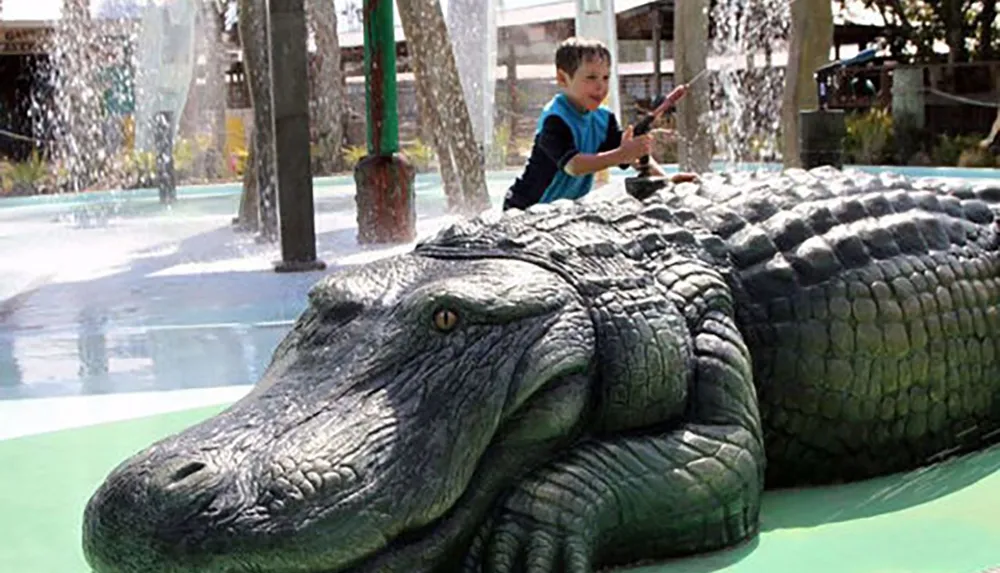 A child is pretending to ride a life-sized crocodile sculpture at a water play area