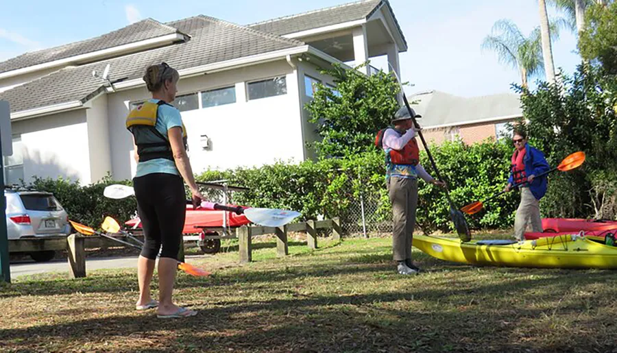 Three people wearing life jackets are preparing or discussing their kayaking trip on a grassy area with two kayaks and paddles visible, in a suburban setting.