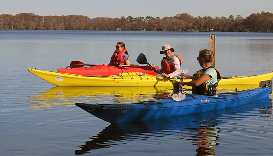 Three individuals in life jackets are kayaking on calm water, with one holding up a paddle in greeting or pose.