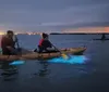 People are kayaking at dusk with illuminated kayaks that cast a blue glow on the water