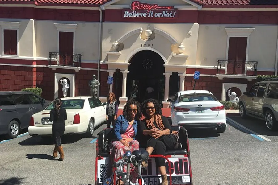 Two individuals are seated on a rickshaw in front of the entrance to Ripley's Believe It or Not! museum, with cars parked nearby and another person walking in the background.