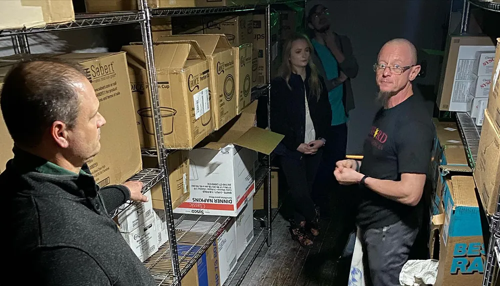 A man in a black T-shirt is engaging with another person in a dimly lit storage area surrounded by cardboard boxes while two other individuals observe