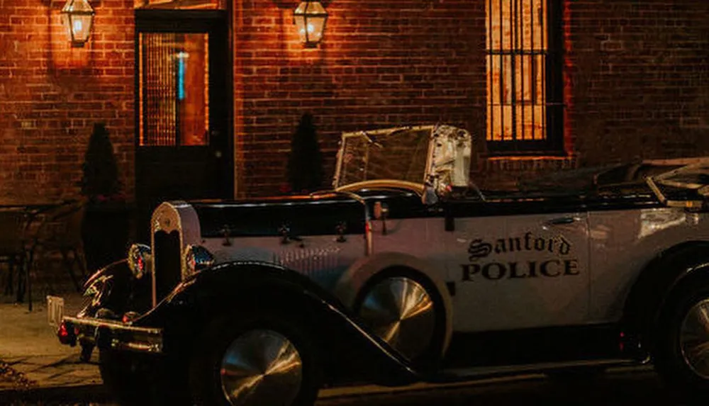 A vintage police car with Sanford Police written on the side is parked by a brick building under warm glowing lights at night