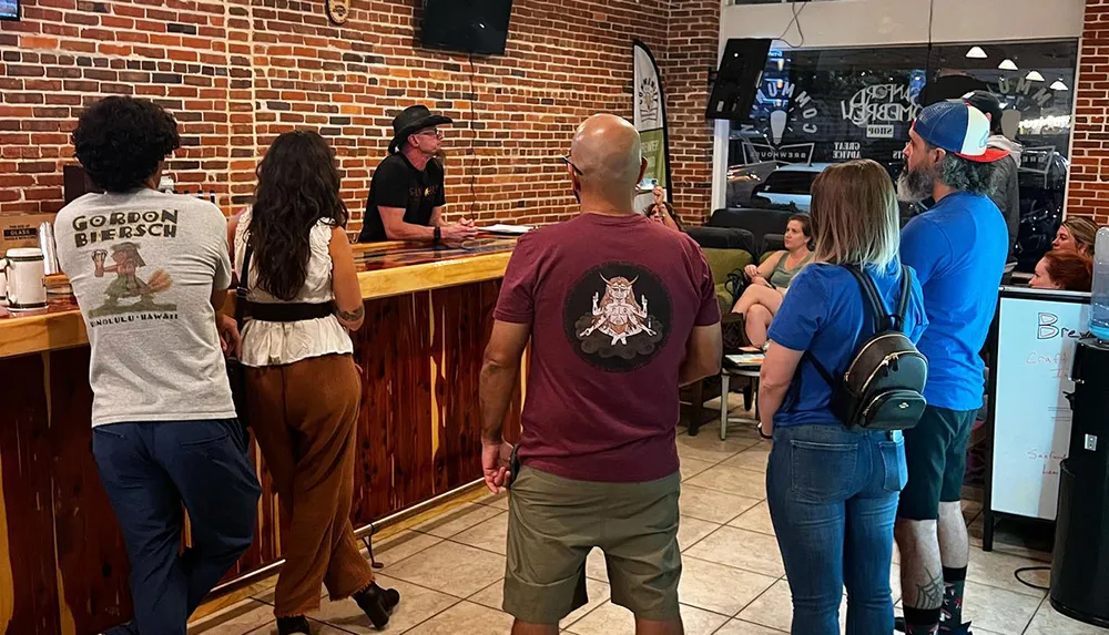 Customers are casually standing and sitting inside a bar with exposed brick walls as they interact with a bartender behind the counter