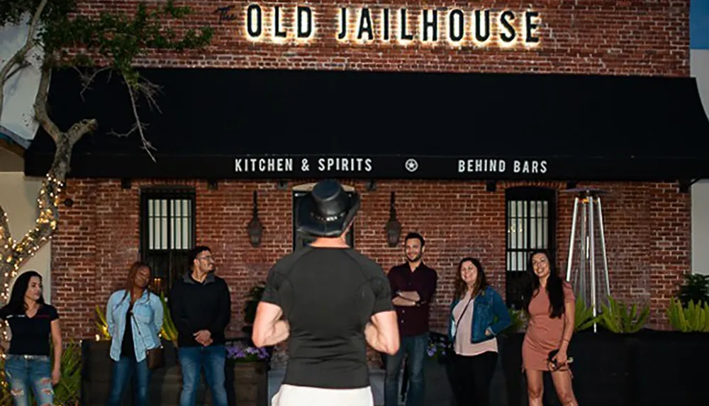 A group of people listen to a person in a hat speaking in front of the OLD JAILHOUSE Kitchen  Spirits Behind Bars restaurant showcasing its unique theme