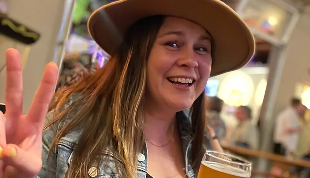 A smiling person with a hat gives a peace sign while holding a glass of beer in a lively indoor setting