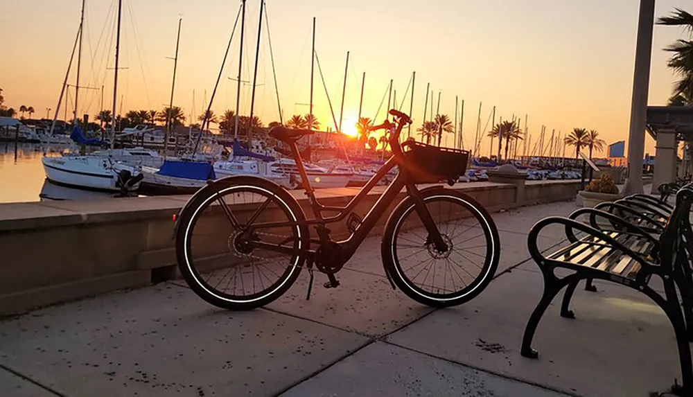 A bicycle is parked along a marina walkway with yachts at sunset creating a silhouette against the vibrant sky