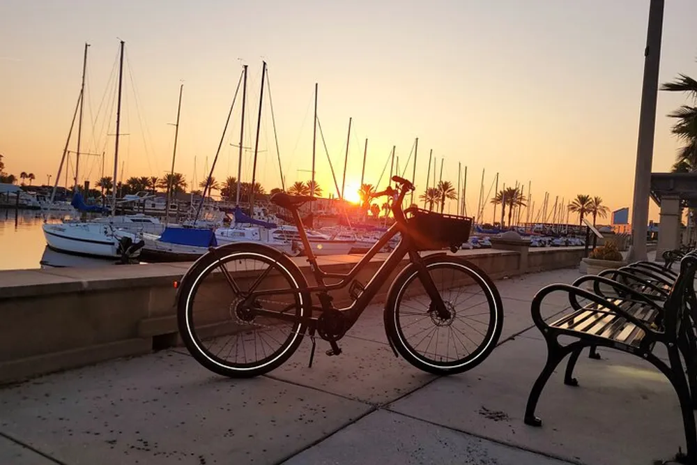 A bicycle is parked on a waterfront promenade with benches overlooking a marina filled with sailboats basking in the warm glow of a setting sun
