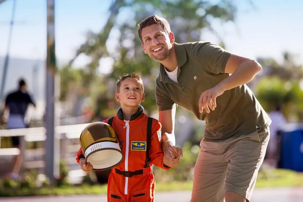 A smiling adult is crouching next to a child dressed as an astronaut both looking up with expressions of anticipation or excitement