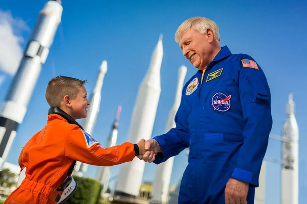 A child dressed as an astronaut shakes hands with an adult astronaut in front of a display of rocket models under a clear sky