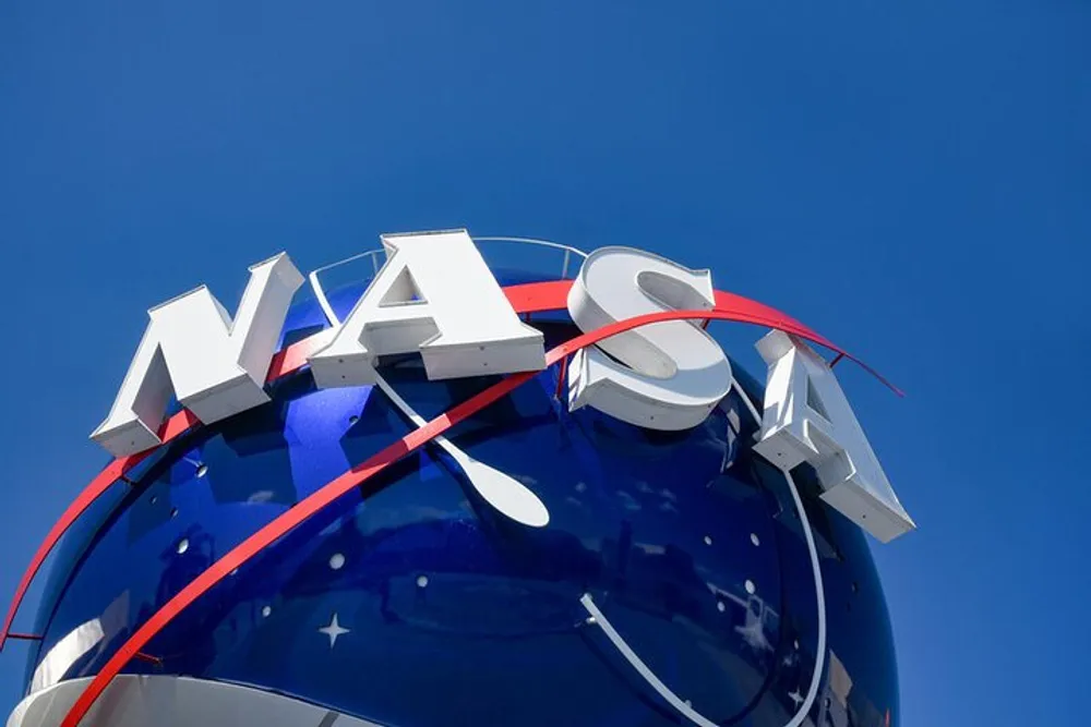 The image shows an upward view of a spherical structure with the NASA logo and a red swoosh against a clear blue sky