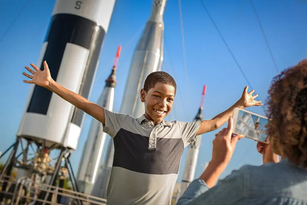 A joyful child spreads his arms wide with a smile while someone takes his photo with towering rockets displayed in the background under a clear blue sky